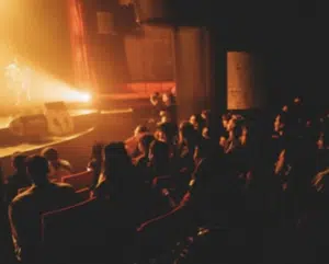 Audience seated in a dimly lit theater watching a brightly lit stage performance.